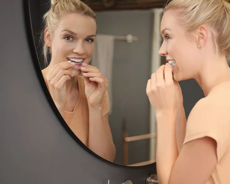 Smiling woman looking in a mirror while inserting clear teeth aligners